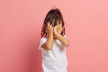 Küçük Asyalı kızın korkuyla yüzünü örten stüdyo portresi, bir şeyden korkuyor, pembe izole edilmiş bir arka planda duruyor. Çocukların korkuları ve fobileri