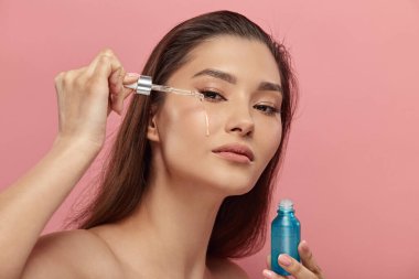 Cazibeli bayan günlük güzellik gösterisi yapıyor. Mavi şişedeki kozmetik ürünle pipette düşürücü kullanıyor. Üstsüz, uzun saçlı bir kadın, yüz yağıyla ideal sağlıklı cilde retinol uyguluyor. Boşluğu kopyala.