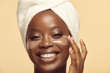 Beyaz kaplıca türbanlı Afro etnik kökenli genç bir kadının portresi sarıda kozmetik yüz tedavisinden zevk alıyor. Mutlu kahverengi tenli kız gözlerinin altına hidrojel bantlar koyar. Güzellik ve kişisel bakım.