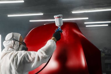 Koruyucu maskeli profesyonel sprey boyacı ve arabanın kaputunu boya odasında sprey tabancayla kırmızıdan kırmızıya boyayan kıyafetler. Boya kabininde araç boyama, restorasyon ve onarım..