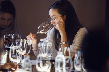 Dişi sommelier dikkatle beyaz şarap aromasını kokluyor, bardağı burnuna dayayıp, çeşitli şarap kadehleri ve su şişeleriyle donatılmış masada otururken, çalışmanın bir parçası olarak
