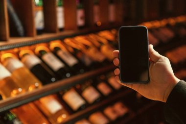 Bir insan, sıcak mahzendeki raflarda bulanık şarap şişelerinin önünde boş ekranlı bir akıllı telefonu tutar..