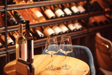 Kırmızı ve beyaz şarap şişeleri yuvarlak ahşap bir masada iki bardak, şarap rafları ve yumuşak ışıklandırma ile çevrili.