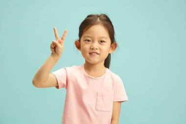 Mutlu ve gülümseyen Çinli ilkokul çocuğu barış jesti gösteriyor, nezaketini ve samimiyetini ifade ediyor, şirin ve hoş bir görünüşü var, tarafsız pembe tişörtü var ve mavinin karşısında duruyor.