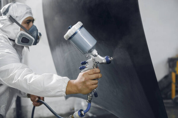 Spray painter worker in protective glove with airbrush pulverizer varnishing and painting car body in paint chamber. Car vehicle part in garage during repair work.