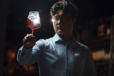 Sommelier, loş ışıklı şarap mahzeninin ortasında dikiliyor, dikkatle kırmızı şarap kadehinin rengini ve berraklığını gözlemliyor, viskozitesini ve renklerini ahşap şarap raflarının arka planında değerlendirmek için camı ustalıkla eğiyor.