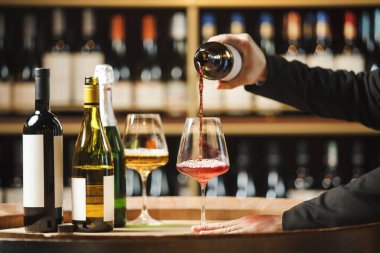 Tanınmayan şarap garsonu, şarap şişeleriyle dolu ahşap raflarla doluyken yeraltı mahzeninde şişeden cama kırmızı şarap dolduruyor..