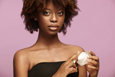 Kusursuz kadın derisi için kare kavanozda krema. Afro-Amerikan modeli kozmetik kutusundan nemlendirici sürüyor. Güzellik ve kişisel bakım, cilt bakımı, kozmetoloji. Mor üzerine ön görünüm.