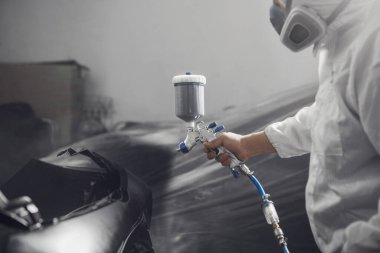 Tamirat sırasında boya odasında sprey tabanca kullanarak arabanın bir kısmını siyaha boyayan otomobil tamircisi. Arabanın onarımı için servis. 