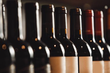 Şarap şişelerinin seçimi ahşap rafta, çeşitli etiketlerin sergilendiği ve şişelerin zarif hatlarının gösterildiği, ön plandaki şişelere odaklanmış, hafif bulanık