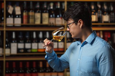 Profesyonel şarap uzmanı, şık mavi gömlekli, düşünceli bir şekilde ayakta duruyor, bir bardak beyaz şarabı sapının yanında tutuyor, buketini nazikçe açmak için şarap şişesini nazikçe çeviriyor.