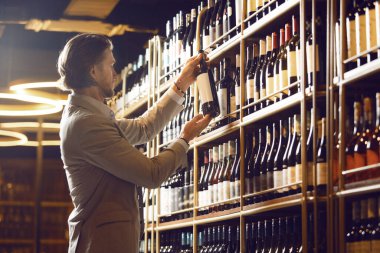 Erkek sommelier şarap şişesini seçiyor, şarap mahzeninde duruyor, büyük kadehlerle dolu şişeler ve nefis alkollü içecekler büyük bir koleksiyona koyuyor, tüm olası zevkleri olan..