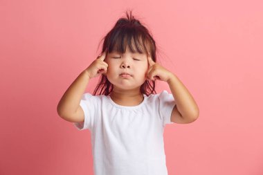 Ciddi küçük Asyalı etnik kızın portresi, önemli bir şeyi hatırlamak için parmaklarını şakaklarda tutar ve gözlerini kapatır. Pembe tişörtün üzerinde tek başına duran beyaz tişört giyer.