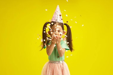 Küçük mutlu Asyalı etnik kızın izole edilmiş stüdyo portresi parti şapkasıyla konfeti üflüyor, doğum gününü ya da başka bir bayramı kutluyor, şenlik kıyafetleri içinde, komik bir ruh hali içinde ayakta duruyor.