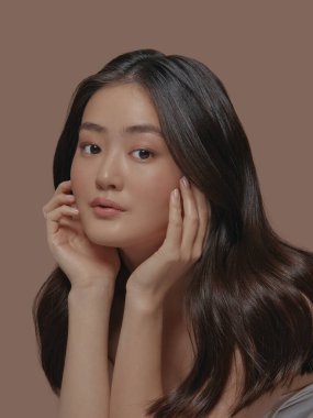 Uzun esmer, çekici, çekici bir Japon kadının portresinin dikey çekimi. Bej bir arka planda tek başına duran yüzüne dokunuyor. Cilt bakımı, kozmetoloji ve plastik