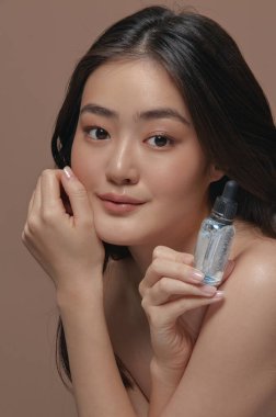 Dikey yakın plan portre, sağlıklı ve pürüzsüz bir cilde sahip Koreli güzel bir kadın bej rengi izole arka plan üzerinde poz veren yüz serumunu tutuyor. Cilt bakımı