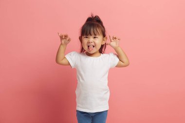 Küçük güzel Asyalı kızın stüdyo portresi eğleniyor ve ellerini pembe izole edilmiş arka planda kaldırarak dilini gösteriyor..
