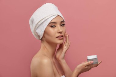 Banyo havlusu giymiş çekici Asyalı kadın, avuç içi boş kozmetik kavanozunda saç sarığı var. Kadın modeli nemlendirici için konteynır gösteriyor. Yüz, vücut kremi ambalajlama.