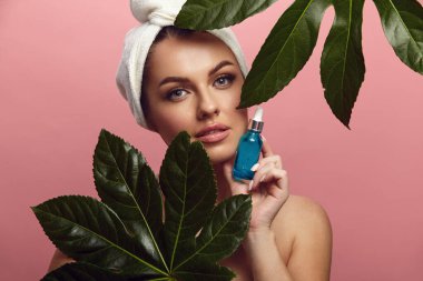 Güzellik görüntüsü, temiz bir cilde sahip genç bir kadın yüz bakımı için nemlendirici serum tutuyor ve pembe izole arka planda tropikal yaprağın yanında duruyor..