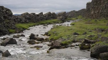 Yavaş çekimde, İzlanda 'daki engebeli lav kayaları ile yeşil çimenli bankalar arasında kıvrılan huzurlu bir nehir görüntüsü elde ediliyor. Yumuşak hareket, akan su ve antik volkanik arazi arasındaki zıtlığı vurgular..
