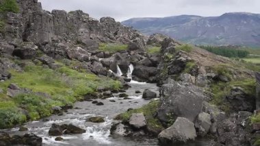 Video İzlanda 'daki karanlık volkanik kayalar arasında akan küçük ama güçlü bir şelale yakalar. Yeşil yosunlar ve engebeli uçurumlarla çevrili sahne İzlanda doğasının saf güzelliğini vurguluyor..