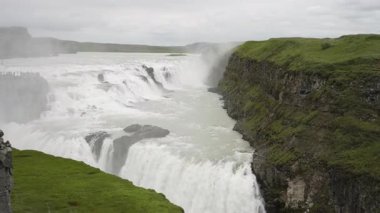 İzlanda 'daki Gullfoss şelalesinin tüm gücünü gösteren video kayalıklardan aşağı süzülürken. Turistler, etrafı sislerle çevrili ve dramatik bir manzarada doğanın kükremesiyle çevrili bir perspektiften izliyorlar..