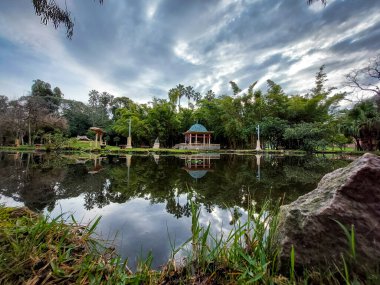 Sakin göle yansıyan doğulu köşkü olan bahçe, ağaçlarla çevrili ve bulutlu gökyüzü sakin, dengeli bir manzara..