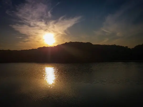 Güneş ormanın üzerinde batıyor, sakin sulara yansıyor, günün sonunda yumuşak bulutlar çerçeveleniyor..