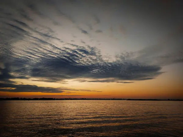 Dalgalı bulutlu gökyüzü alacakaranlıkta, suyun üzerinde altın yansıması ve ince bir ufuk çizgisi ile.