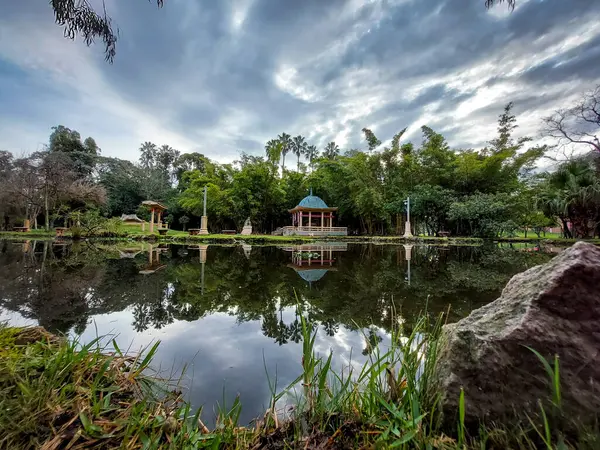 Sakin göle yansıyan doğulu köşkü olan bahçe, ağaçlarla çevrili ve bulutlu gökyüzü sakin, dengeli bir manzara..