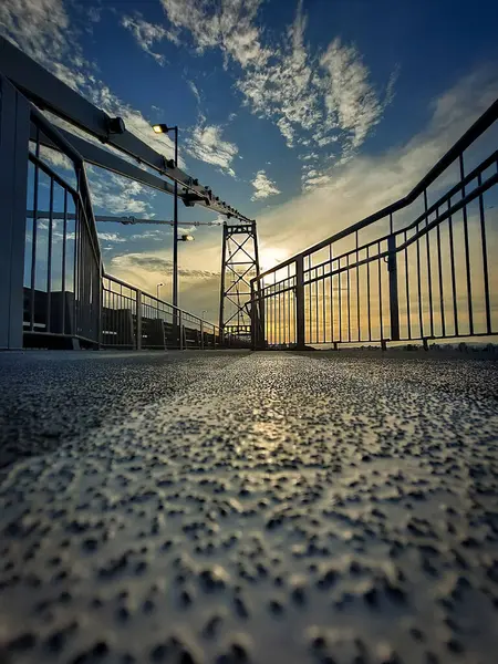 Zemin kattan görünen metal köprü, odak noktasında dokulu asfalt, çarpıcı bir gökyüzü altında demiryolu ve günbatımı ışığı ile çevrili..