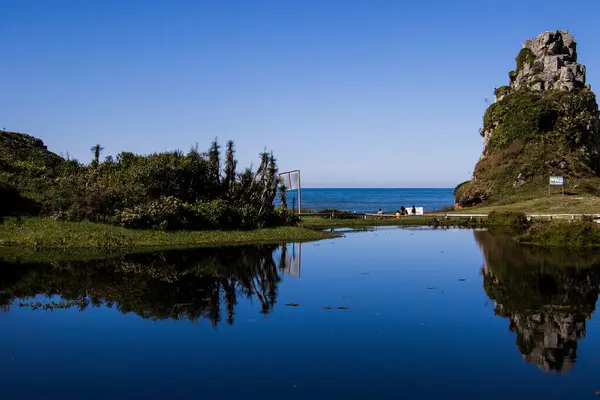 Kaya oluşumu ve yoğun bitki örtüsü, okyanus manzaralı ve mavi gökyüzüyle sakin bir göle yansıyor..