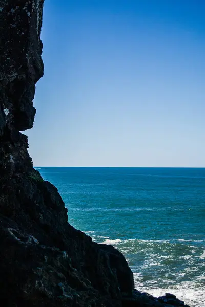 Görüntü ön planda koyu kayalık bir uçurum ve arka planda açık bir gökyüzünün altında mavi bir okyanus gösteriyor..
