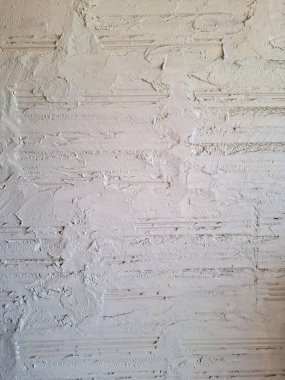 Beyaz alçıyla kaplanmış, desenli yüzeyleri, yatay tuğla çizgileri ve alçıdaki kusurları gösteren, kabartma ve lekeli boya bantlarıyla kaplı bir duvarın yakın plan görüntüsü..