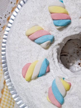 Yüzük şeklinde bir kekin yakın plan görüntüsü üzerinde beyaz bir hindistan cevizi tabakası var. Üst kenarı etrafında düzenli aralıklarla, kat kat, çarpık pastel renkli marşmelovlar; pembe, sarı ve mavi; düzenlenir. Karşıtlık olsun.