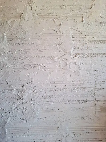Beyaz alçıyla kaplanmış, desenli yüzeyleri, yatay tuğla çizgileri ve alçıdaki kusurları gösteren, kabartma ve lekeli boya bantlarıyla kaplı bir duvarın yakın plan görüntüsü..