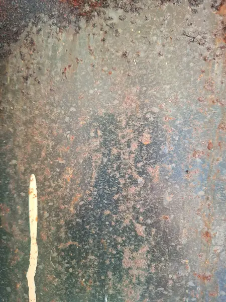Eski metal bir yüzeyin yakın plan görüntüsü aşınma ve dağınık pas lekeleri gösteriyor. Tabanda gri ve yeşilimsi renkler bulunurken, küçük benekler ve yıpranmış metal alanlar yıpranmış görünümü vurgular. Sol kenarda, bir dikey d