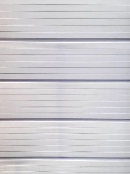 Yatay panellerden oluşan beyaz boyanmış metal bir garaj kapısının yakın plan görüntüsü. İnce kanallarla birleştirilmiş. Yüzey yumuşak ışık yansımaları ve panel eklemlerinde yumuşak gölgeleme ile pürüzsüz bir doku sergiliyor. Tekrarlanan yatay 