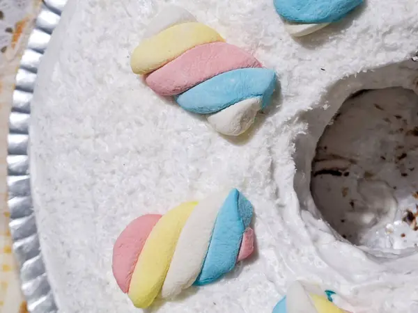 Görüntü, yüzeye tanecikli bir doku veren, parçalanmış beyaz hindistan cevizi tabakasıyla kaplı halka şeklinde bir kekin yakın çekim görüntüsünü sunuyor. Bükülmüş pastel renkli şekerlemeler (pembe, sarı, mavi ve beyaz) simetrik olarak c 'nin üzerine yerleştirilir.