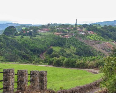 Yeşil tepeler, yamaçlara dağılmış evler, yoğun bitki örtüsü ve hafif bulutlu bir gökyüzü içeren yemyeşil bir kırsal manzaranın panoramik görüntüsü. Huzurlu ve huzurlu bir ortam. Doğayla barışı ve bağı çağrıştırıyor..