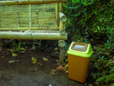 Açık hava ortamında bambu bir bankın yanına yerleştirilmiş sarı bir çöp tenekesinin fotoğrafı. Endonezya veya Güneydoğu Asya 'da temizlik, çevre bilinci ve sürdürülebilir yaşam tarzını teşvik eder.