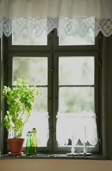 Köy evindeki pencere eşiğinde mumlar, cam şişeler ve kahverengi saksıda bir saksı var..