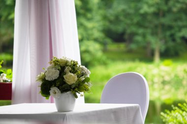 Bir buket beyaz gül ve karanfil bir masanın üzerinde beyaz bir masa örtüsüyle, bir sandalyenin yanında, yemyeşil bir arka planda duruyor.