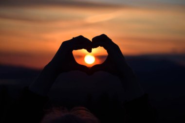 Bir tarlanın ortasında gün batımından kalp yapan bir kadının silüeti.