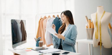 Asyalı genç kadın moda tasarımcılığının ofisinde ve elinde tablet, dizüstü bilgisayar ve akıllı telefon var.
