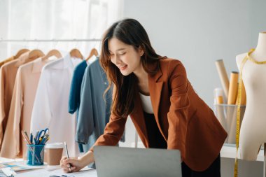 Başarılı bir moda tasarımcısı. Atölyede dururken gülümseyen çekici Asyalı kadın. Güzel iş kadını çalışıyor ve stüdyoda tablet, laptop ve akıllı telefonu tutuyor..