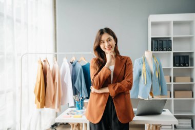 Başarılı bir moda tasarımcısı. Atölyede dururken gülümseyen çekici Asyalı kadın. Güzel iş kadını çalışıyor ve stüdyoda tablet, laptop ve akıllı telefonu tutuyor..