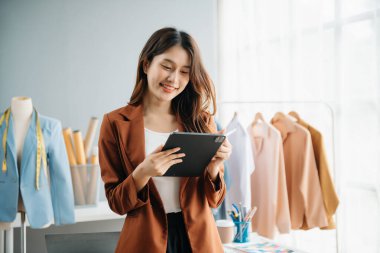 Başarılı bir moda tasarımcısı. Atölyede dururken gülümseyen çekici Asyalı kadın. Güzel iş kadını çalışıyor ve stüdyoda tablet, laptop ve akıllı telefonu tutuyor..