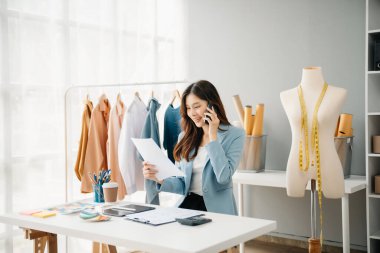 Moda tasarımcısı kadın akıllı telefonlardan konuşuyor ve modern stüdyoda dijital tablet bilgisayarlı dizüstü bilgisayar kullanıyor.