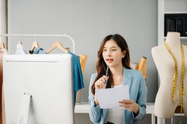 Asyalı genç kadın moda tasarımcılığının ofisinde ve elinde tablet, dizüstü bilgisayar ve akıllı telefon var.